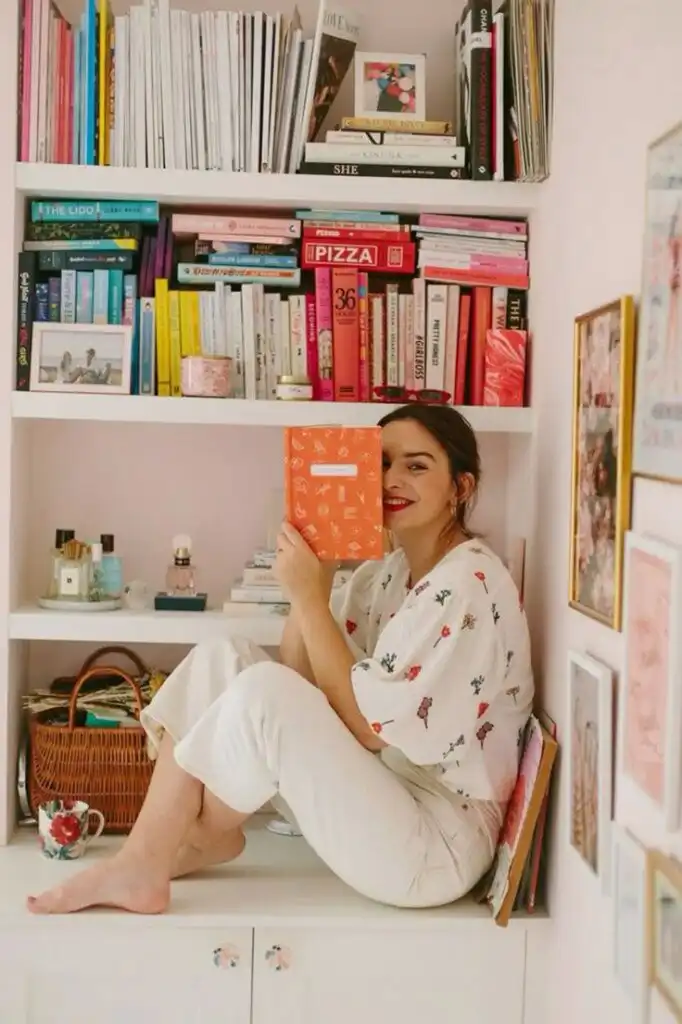 Woman sitting on a bookshelf, holding an orange book, surrounded by colorful books and decor, creating a cozy reading nook.