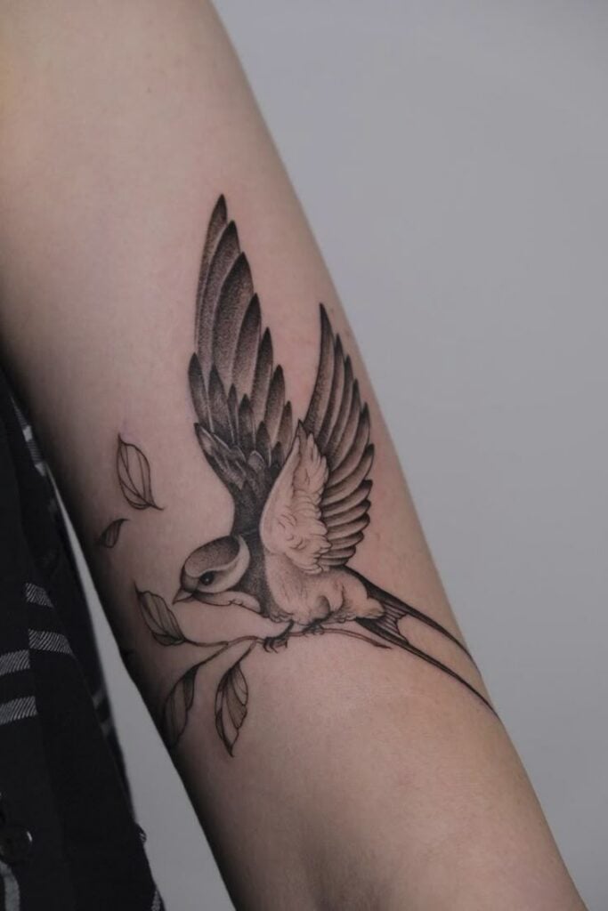 Detailed black and gray bird tattoo with leaves on an arm, showcasing intricate wing and feather design.