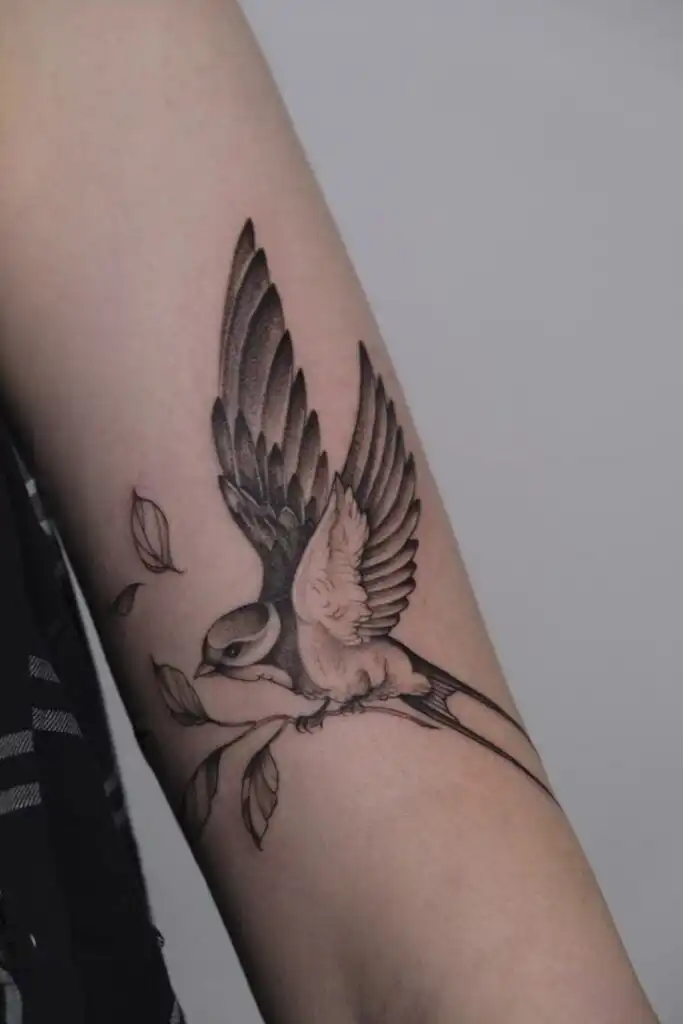 Detailed black and gray bird tattoo with leaves on an arm, showcasing intricate wing and feather design.