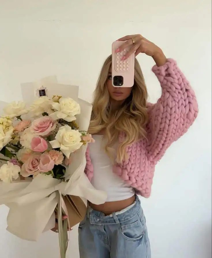 Woman in pink knit sweater holds smartphone and bouquet of pink and white flowers, posing stylishly for a selfie.