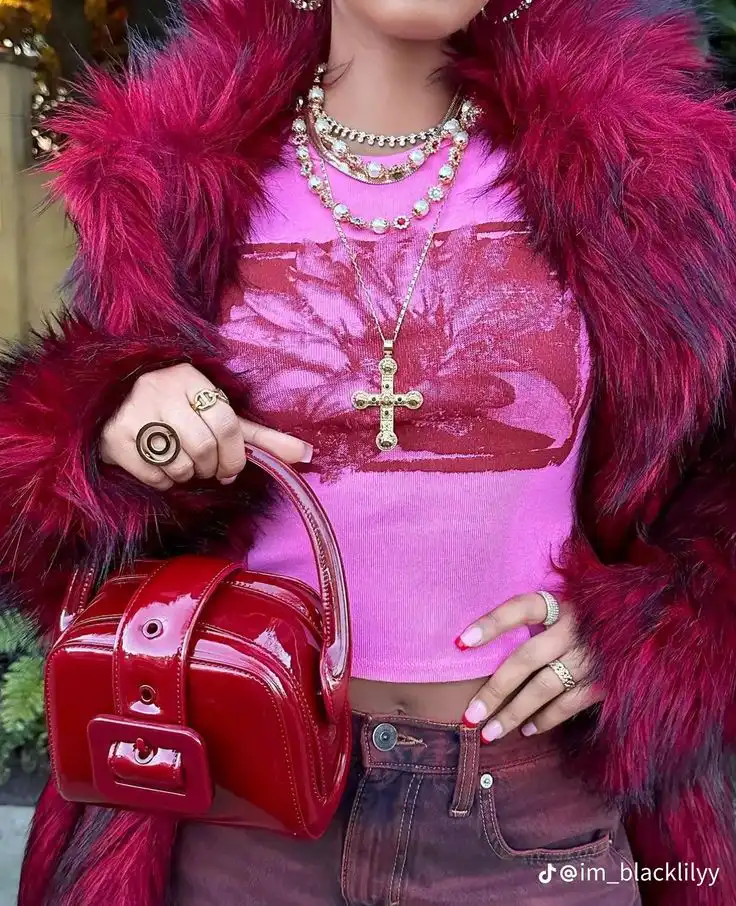 Fashionable woman in pink top and fur coat, accessorized with layered necklaces, rings, and a stylish red patent leather handbag.