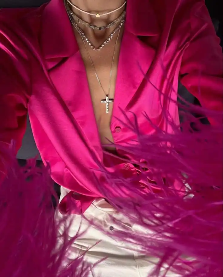 Person wearing a vibrant pink satin blouse with layered necklaces, including a cross pendant, and white pants, surrounded by pink feathers.