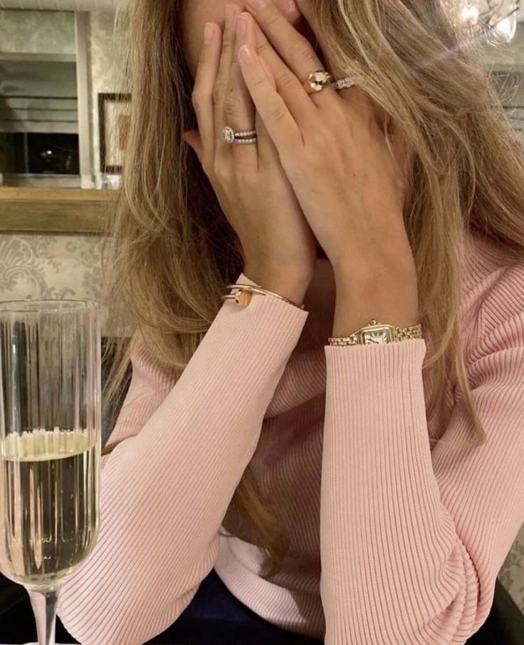 _ 46 Woman with jewelry covers face in pink top, sitting by a champagne glass, conveying emotion or surprise in elegant setting. | Sky Rye Design Woman with jewelry covers face in pink top, sitting by a champagne glass, conveying emotion or surprise in elegant setting.