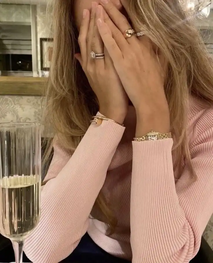 Woman with jewelry covers face in pink top, sitting by a champagne glass, conveying emotion or surprise in elegant setting.