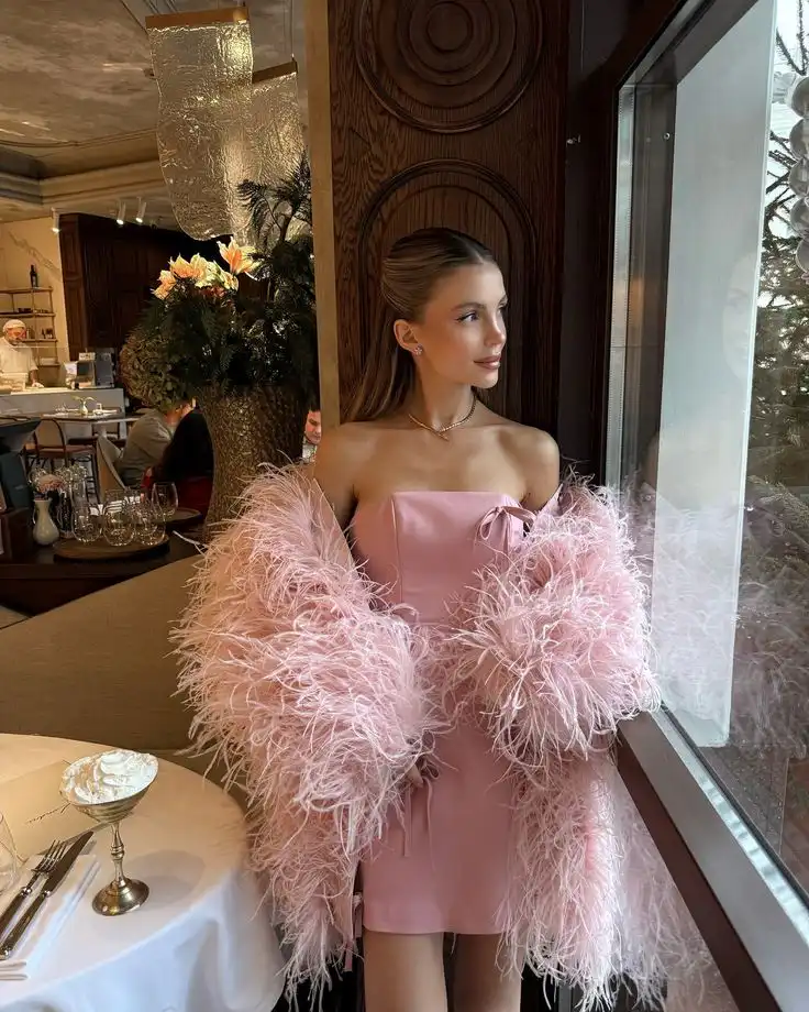 Elegant woman in a pink dress and feather shawl, gazing outside a restaurant window, with a beautifully set table nearby.