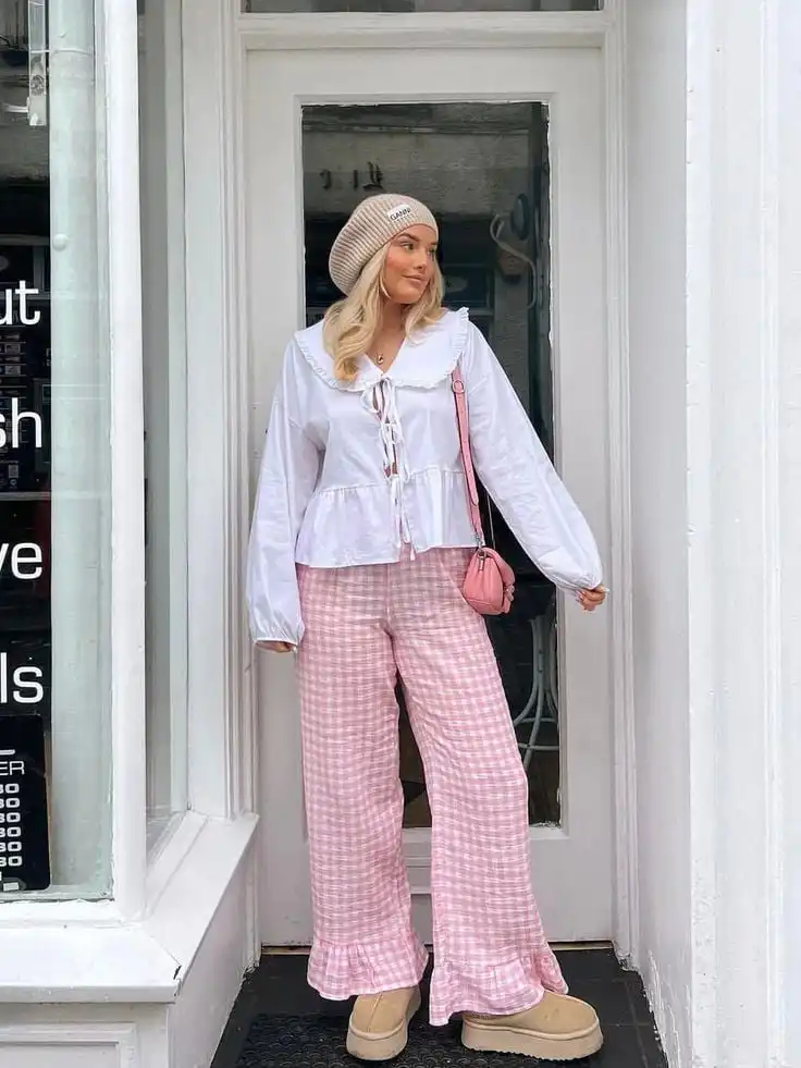 Woman in stylish pink checkered pants, white blouse, and beige beanie poses outdoors with a pink purse. Cozy and fashionable street style.