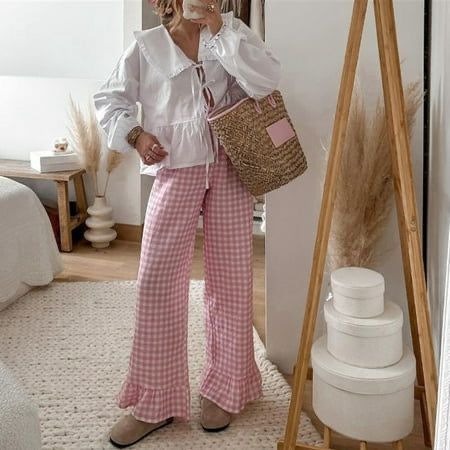 _ 51 Woman in a white blouse and pink checkered pants holds a woven bag in a stylish, cozy bedroom setting with modern decor. | Sky Rye Design Woman in a white blouse and pink checkered pants holds a woven bag in a stylish, cozy bedroom setting with modern decor.