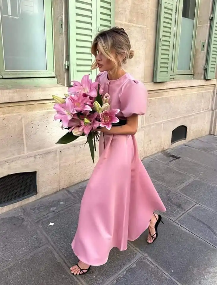 Woman in a flowing pink dress walks with a bouquet of lilies on a chic city street, near pastel green shutters.
