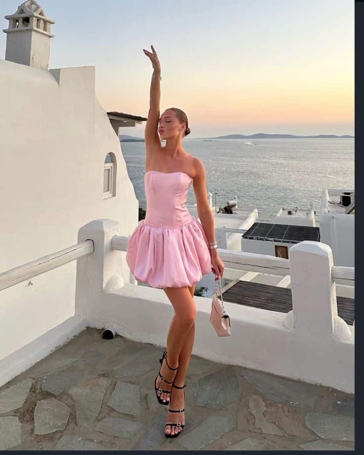 _ 53 Woman in a pink dress poses at a scenic sunset viewpoint overlooking the sea, with white architecture in the background. | Sky Rye Design Woman in a pink dress poses at a scenic sunset viewpoint overlooking the sea, with white architecture in the background.