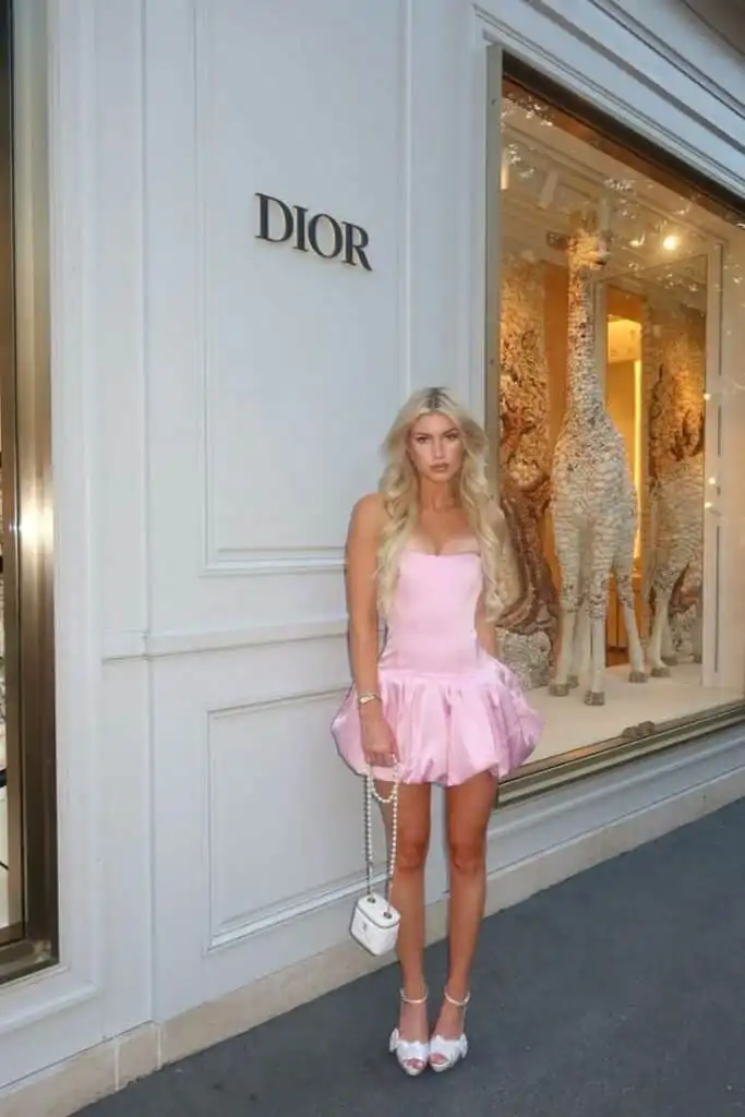 Woman in a pink dress poses stylishly outside a Dior store, holding a small white handbag, with giraffe figures inside the window display.