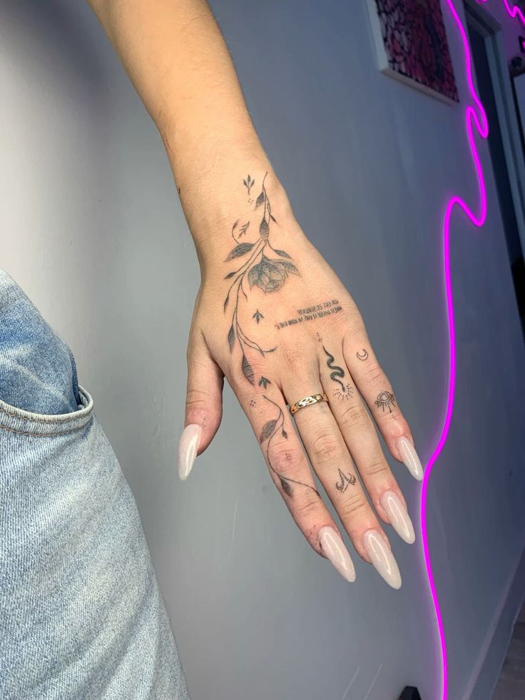 58704431-802e-46bd-b8a7-2231fda18815 Hand with intricate floral and symbol tattoos, wearing nude nails and a gold ring, against a backdrop with pink neon lighting. | Sky Rye Design Hand with intricate floral and symbol tattoos, wearing nude nails and a gold ring, against a backdrop with pink neon lighting.