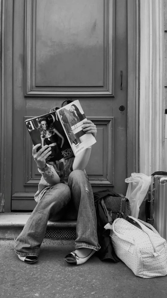 5a708748-fdb4-4cae-b2f0-eba673bcfc8c Person sitting on doorstep reading a fashion magazine, surrounded by bags, suitcase, wearing jeans and ballet flats. Black and white image. | Sky Rye Design Person sitting on doorstep reading a fashion magazine, surrounded by bags, suitcase, wearing jeans and ballet flats. Black and white image.