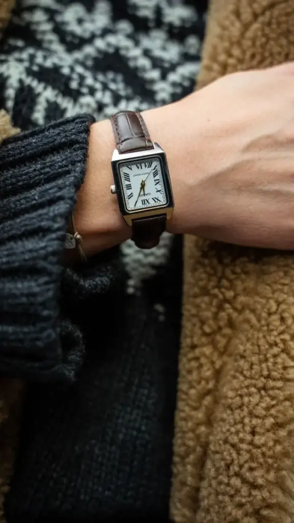 Close-up of a wrist with a brown leather-strap watch, roman numeral dial, and sweater and coat sleeves.