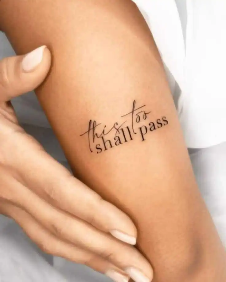 Arm with a tattoo saying this too shall pass in elegant script, gently touched by a hand with nude nail polish, conveying resilience.