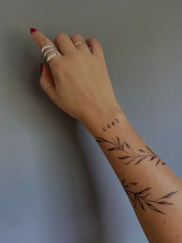 Arm adorned with minimalist tattoo designs, featuring leafy branches and the year 1993, complemented by elegant rings on the hand.