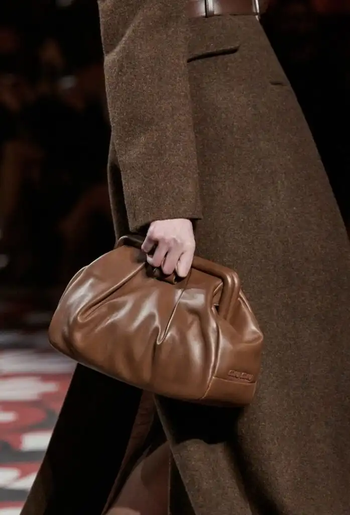 Fashionable brown leather handbag with pleated design, elegantly held by a model in a textured brown coat on the runway.