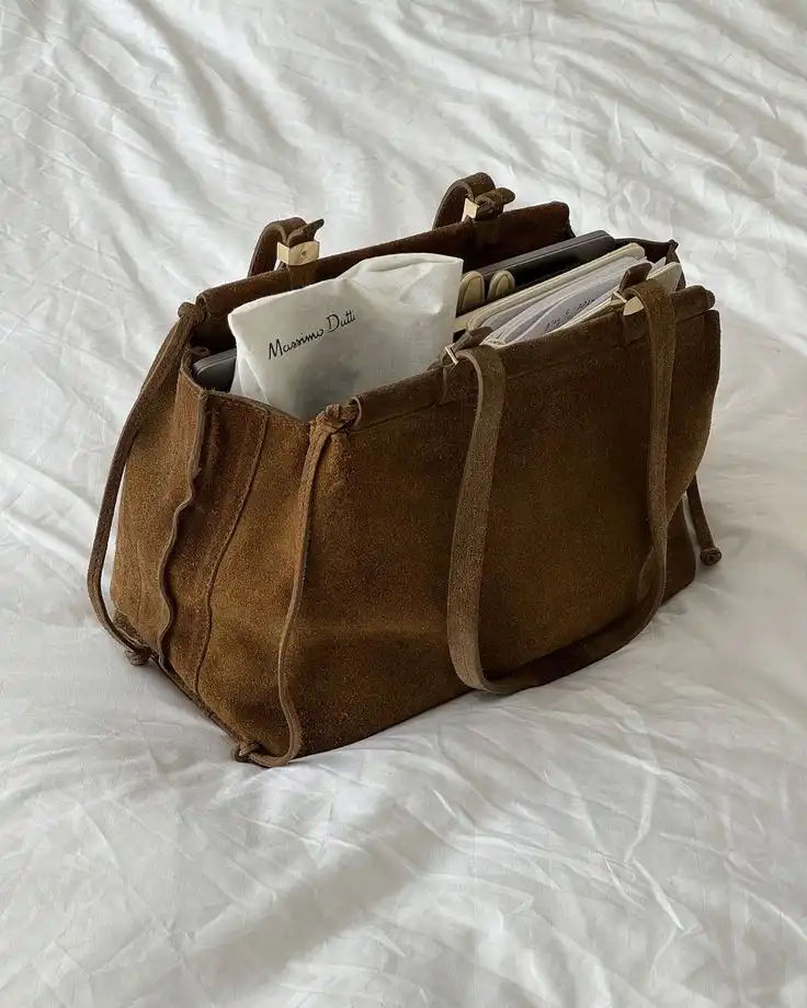 Brown suede bag on white bedding, filled with books and a Massimo Dutti item.