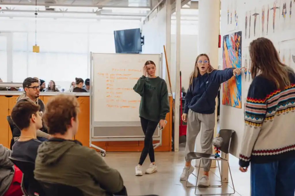 Students engaged in a creative class discussion, presenting artwork and ideas, with a whiteboard displaying notes in a modern classroom setting.