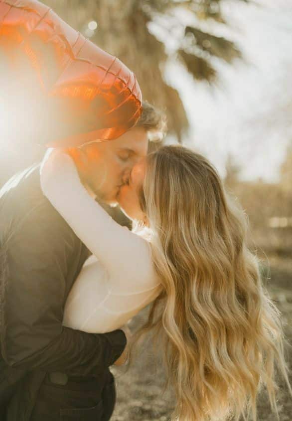 _ 8 Romantic couple kissing outdoors under red heart balloon in soft sunlight, expressing love and affection in a serene setting. | Sky Rye Design Romantic couple kissing outdoors under red heart balloon in soft sunlight, expressing love and affection in a serene setting.