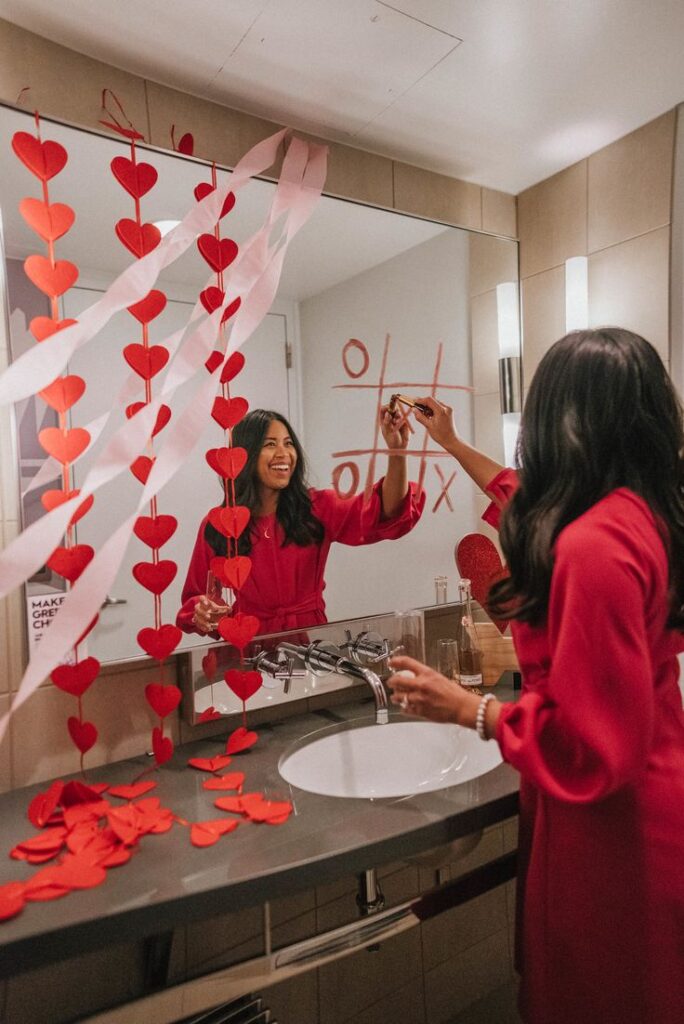 8 Easy Valentines Day Posing Ideas Woman in red dress playing tic-tac-toe on a mirror with lipstick, surrounded by heart decorations in a bathroom. | Sky Rye Design Woman in red dress playing tic-tac-toe on a mirror with lipstick, surrounded by heart decorations in a bathroom.