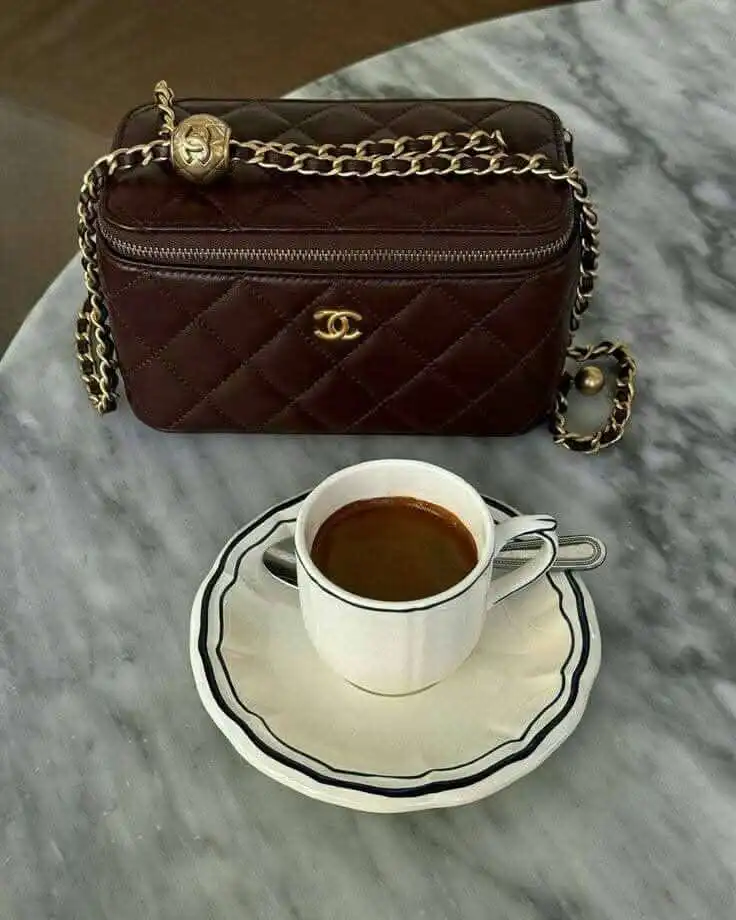Elegant brown quilted handbag with gold chain beside a cup of coffee on a marble table. Cozy luxury fashion and lifestyle.