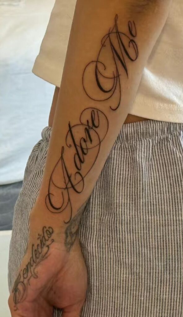 81016215-df03-4efe-834b-375a0e596735 Elegant Adore Me tattoo in cursive on a person's forearm, with Bendecido tattoo in script on the hand, wearing striped pants. | Sky Rye Design Elegant Adore Me tattoo in cursive on a person's forearm, with Bendecido tattoo in script on the hand, wearing striped pants.