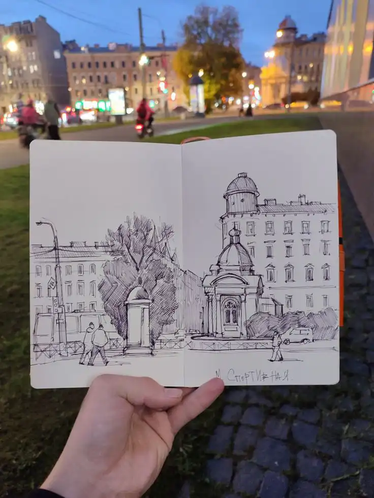 Open sketchbook with a hand-drawn cityscape, reflecting the vibrant urban night scene in the background.