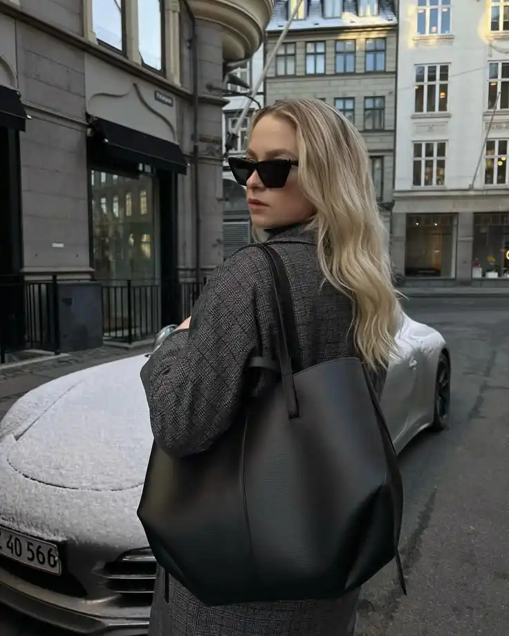 Blonde woman in sunglasses with a black bag, standing in a city street beside a parked car on a snowy day.