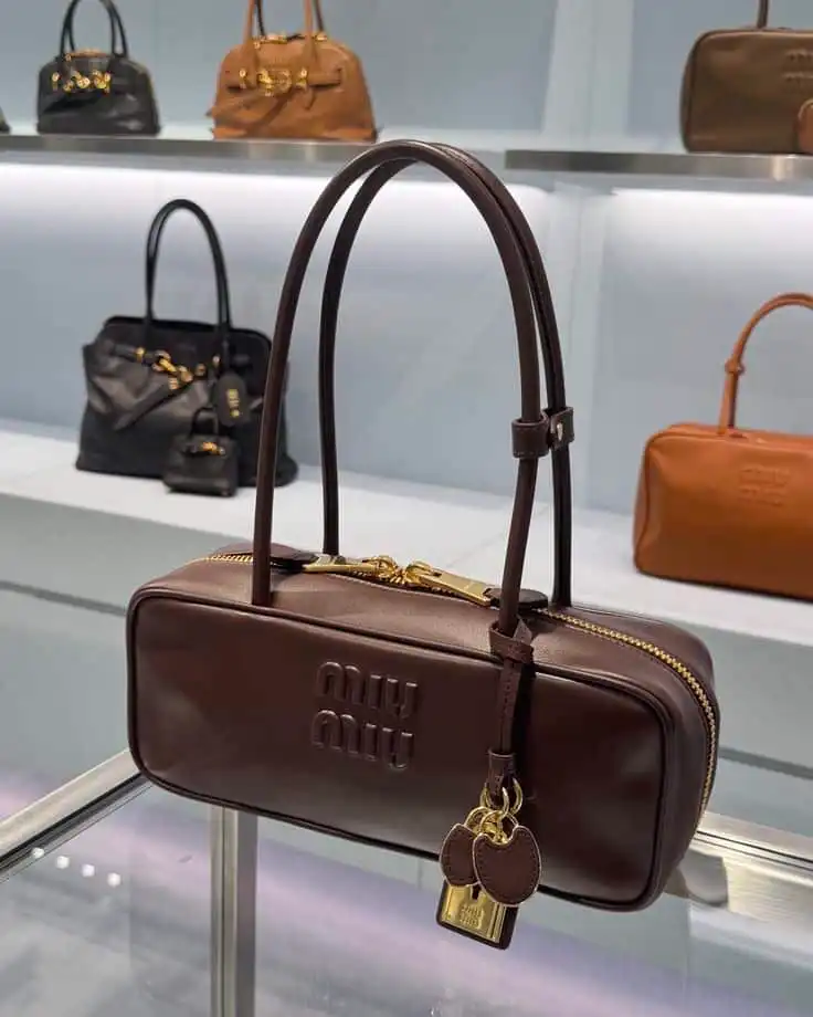 Brown Miu Miu handbag on display in a luxury store, featuring gold accents and keychain, with other bags in the background.