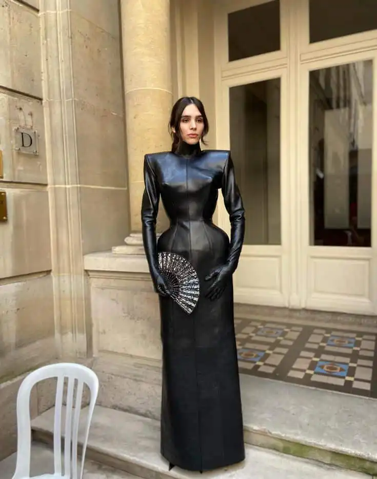 Elegant woman in a long black leather dress, holding a decorative fan, standing in front of a classic building entrance.