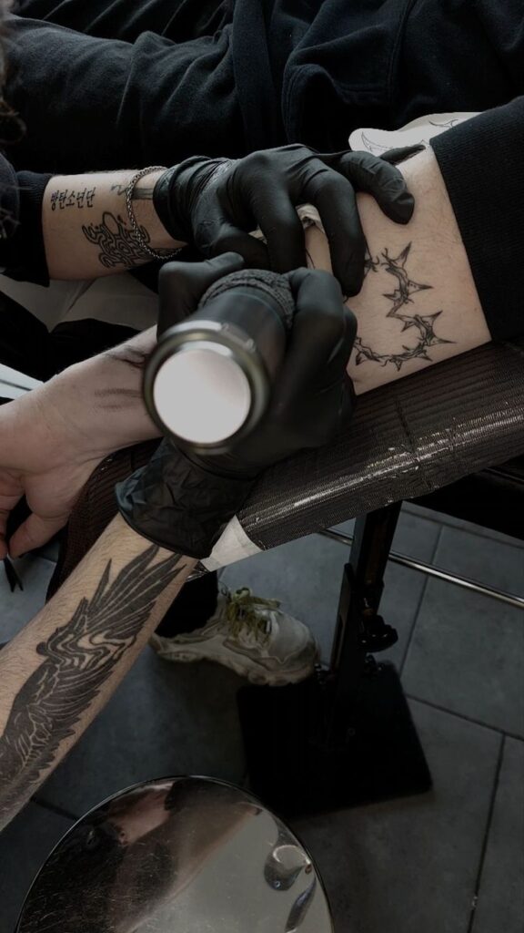 94f46cb0-7314-4911-908e-e4fcb41fddb1 Tattoo artist in black gloves working on forearm design in studio; intricate bat design with detailed linework visible. | Sky Rye Design Tattoo artist in black gloves working on forearm design in studio; intricate bat design with detailed linework visible.