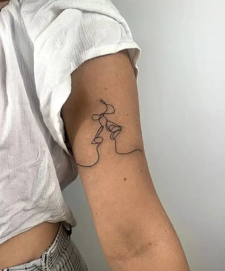 Minimalist one-line tattoo of two faces on a person's upper arm, wearing a white shirt.