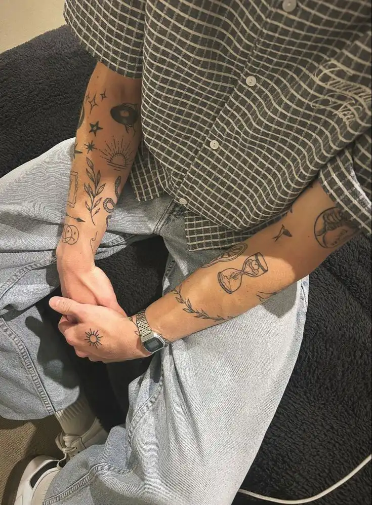 Person with intricate tattoos on arms, wearing a checkered shirt and jeans, sitting with hands folded. Casual style, artistic ink designs.
