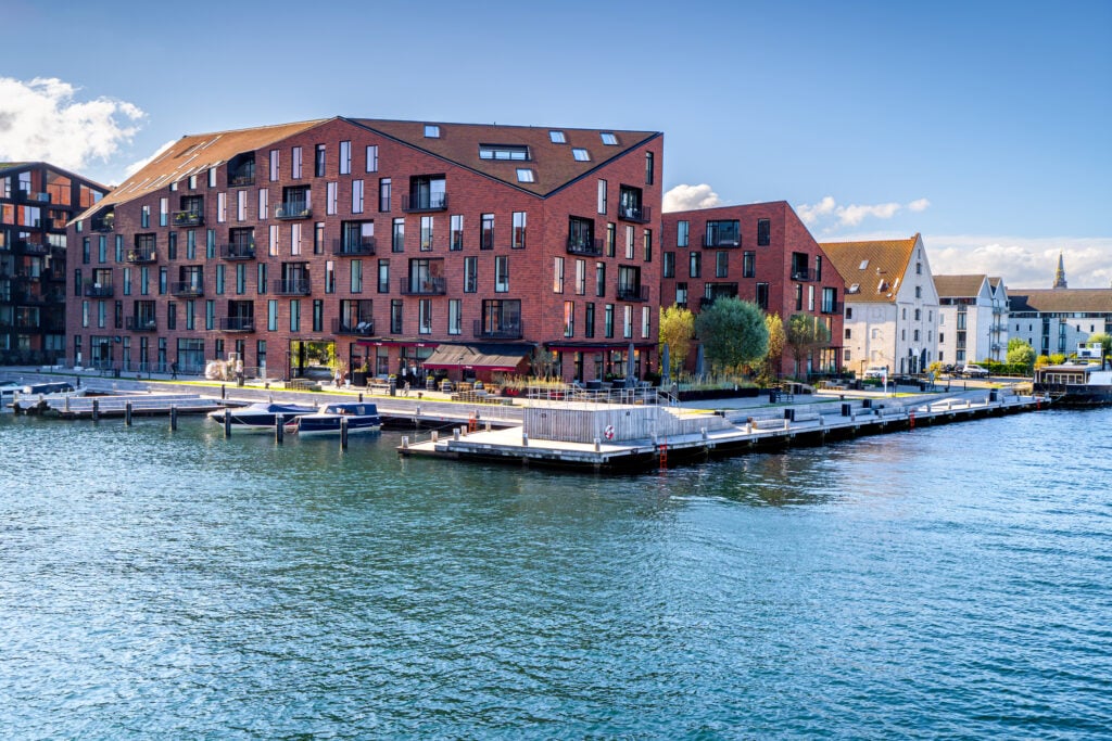 Modern waterfront buildings and dock on a sunny day, featuring a red-brick structure and boats in a calm marina setting.