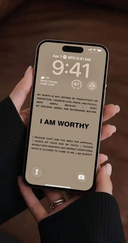 Person holding a smartphone displaying the time, weather, and a self-affirmation message: I am worthy on the screen.