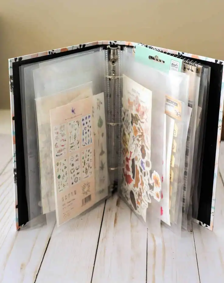 Binder with assorted decorative stickers in plastic sleeves on a wooden table, ideal for scrapbooking and crafting enthusiasts.