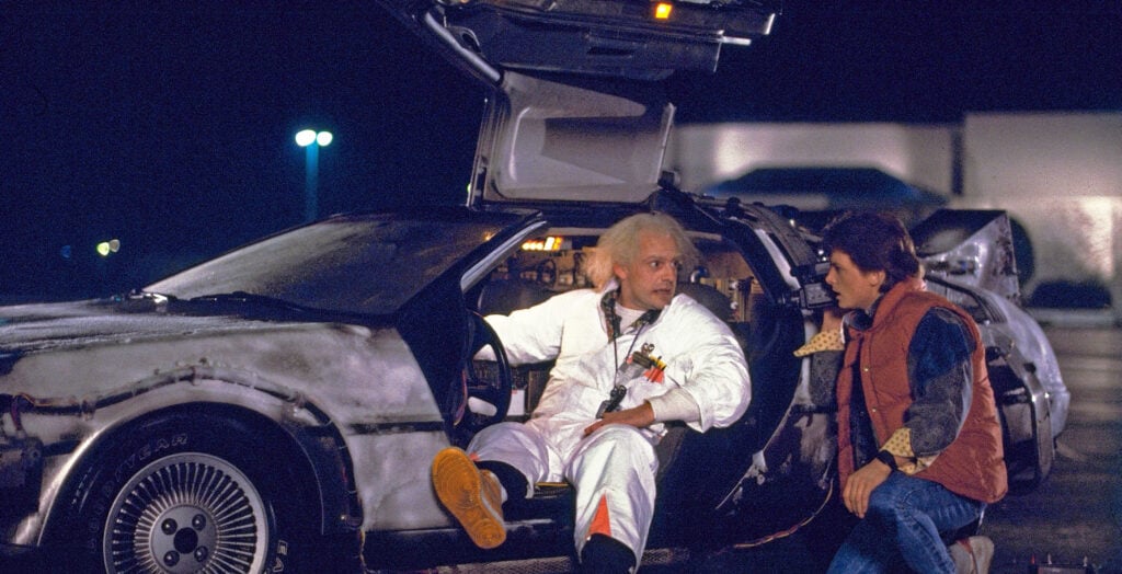 Two characters discuss in front of a futuristic car with gullwing doors at night, one sitting inside while the other listens intently outside.