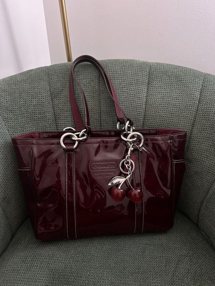 Bags dropping this week 🤍🎀__Available to purchase on 8_14 3_30pm PST ♡__────୨ৎ────_coach coachbag vintagecoach y2k summerbag summertrends bagcollection Maroon patent leather tote bag with cherry charm, resting on a green armchair. Stylish accessory for daily use or special occasions. | Sky Rye Design Maroon patent leather tote bag with cherry charm, resting on a green armchair. Stylish accessory for daily use or special occasions.