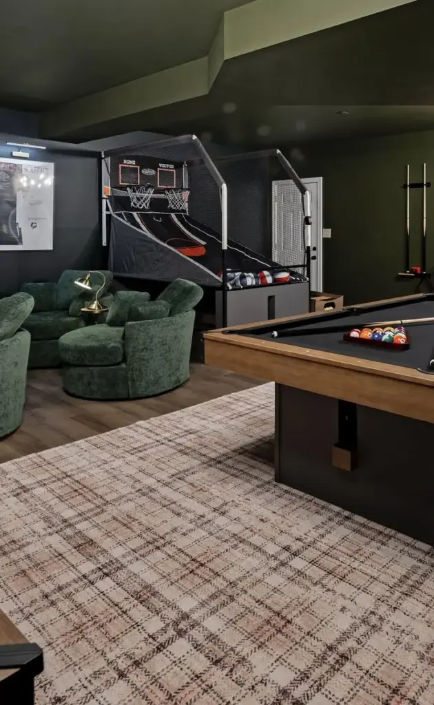 Stylish game room with pool table, basketball arcade game, plush green swivel chairs, and decorative rug for entertainment and relaxation.