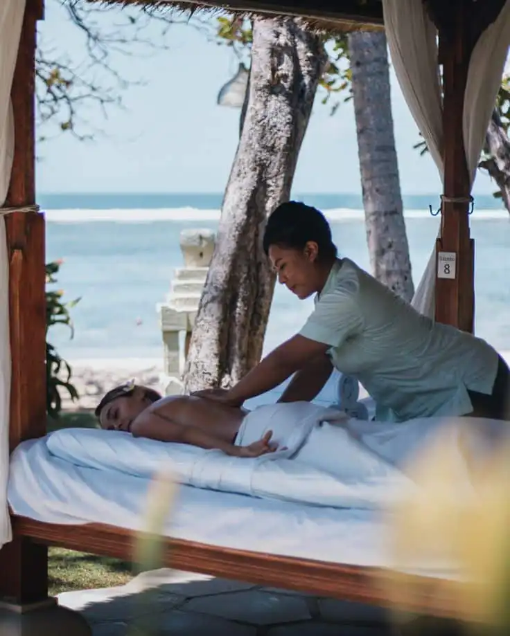 Outdoor massage therapy session by the beach, featuring a relaxing woman on a spa bed under a gazebo with ocean view in the background.