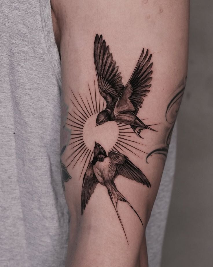 Tattoo of two swallows in flight with sunrays on an arm, symbolizing freedom and love.