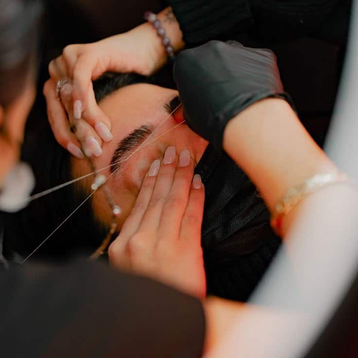 Benefits of Eyebrow Threading_ Dont Dread the_ Person receiving eyebrow threading at a salon, close-up on hands using thread, enhancing facial appearance with precise grooming. | Sky Rye Design Person receiving eyebrow threading at a salon, close-up on hands using thread, enhancing facial appearance with precise grooming.