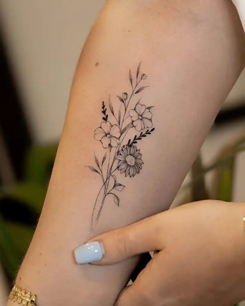 Elegant black floral tattoo on forearm, featuring daisies and leaves, captured with manicured hand for delicate body art inspiration.
