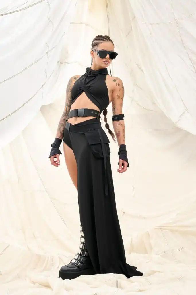 Fashion-forward person in black asymmetric outfit with tattoos and sunglasses, standing confidently against a white draped