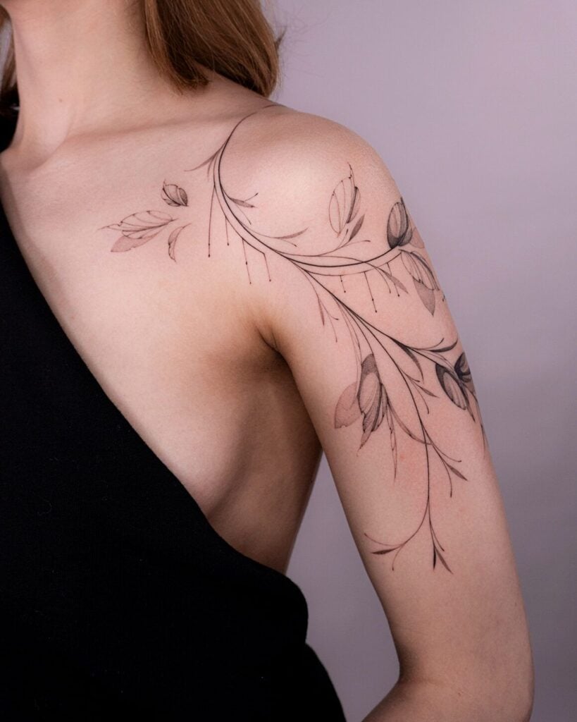 Delicate floral tattoo on woman's shoulder, intricate design with leaves and petals. Black ink art, sleeveless black top.
