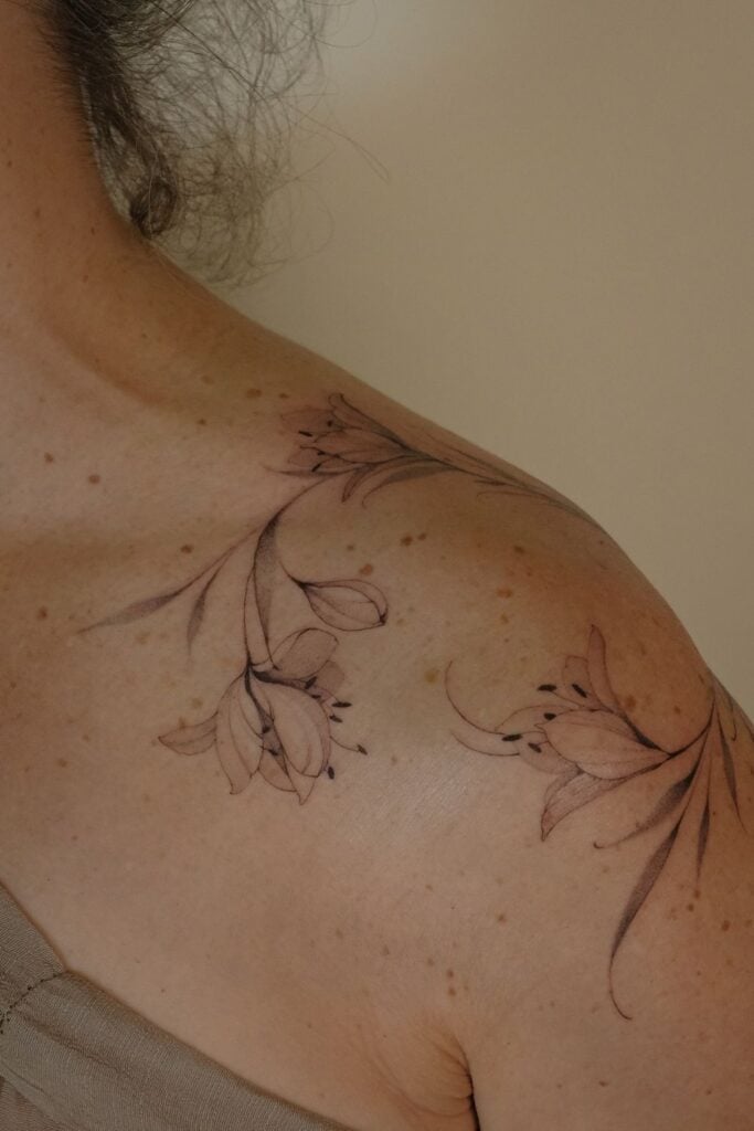 Delicate floral tattoo on a person's shoulder, featuring elegant lily-like flowers and flowing vines on beige background.
