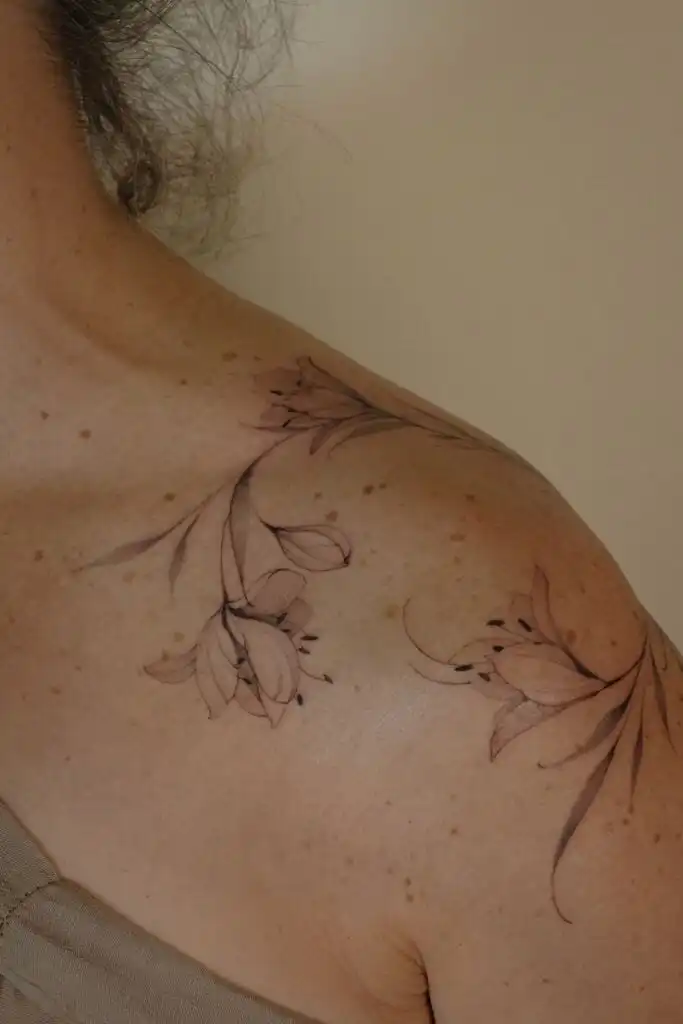 Delicate floral tattoo on a person's shoulder, featuring elegant lily-like flowers and flowing vines on beige background.