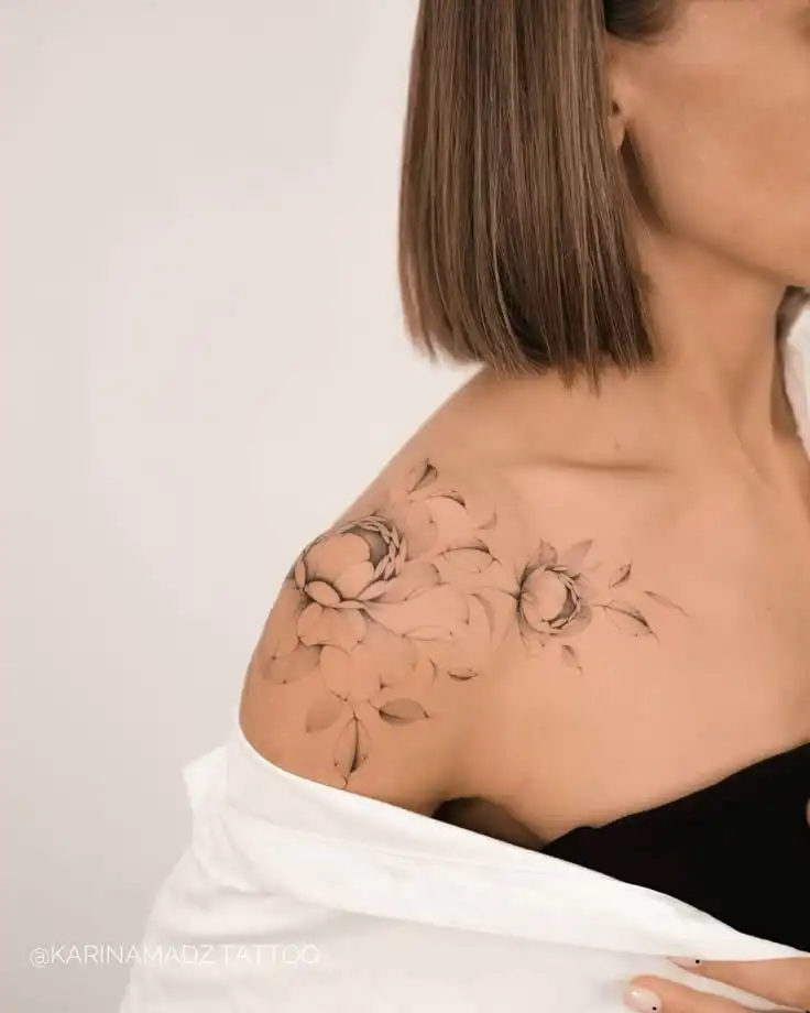 Floral shoulder tattoo on woman with short brown hair, showcasing delicate peonies and leaves, against a neutral background.