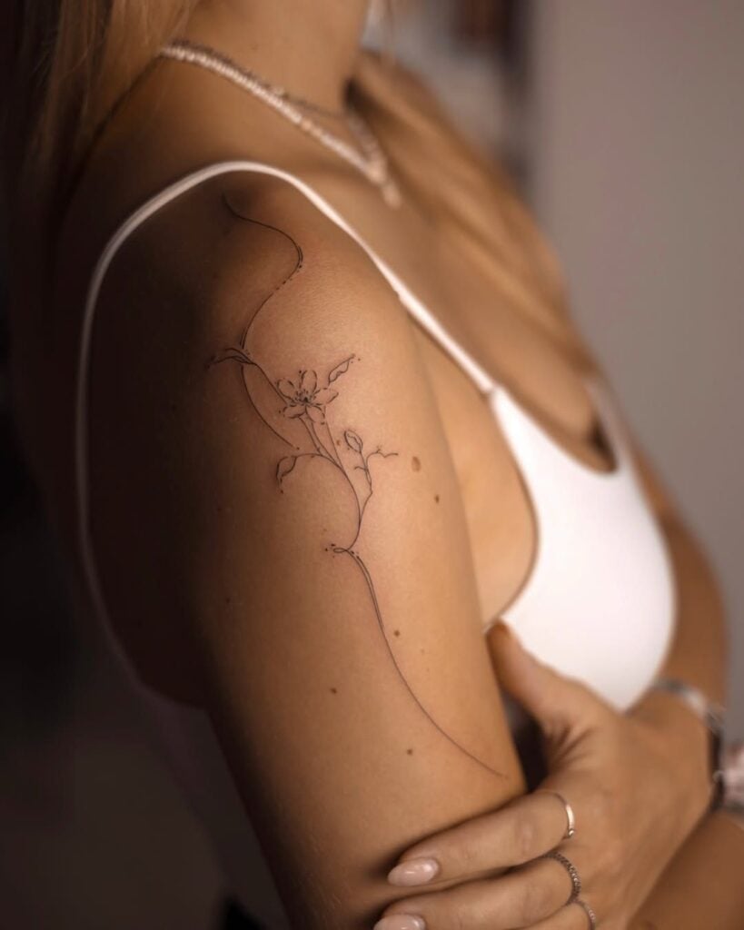 Minimalist floral tattoo on upper arm, woman in white tank top showing delicate line art with a curved stem and flower.