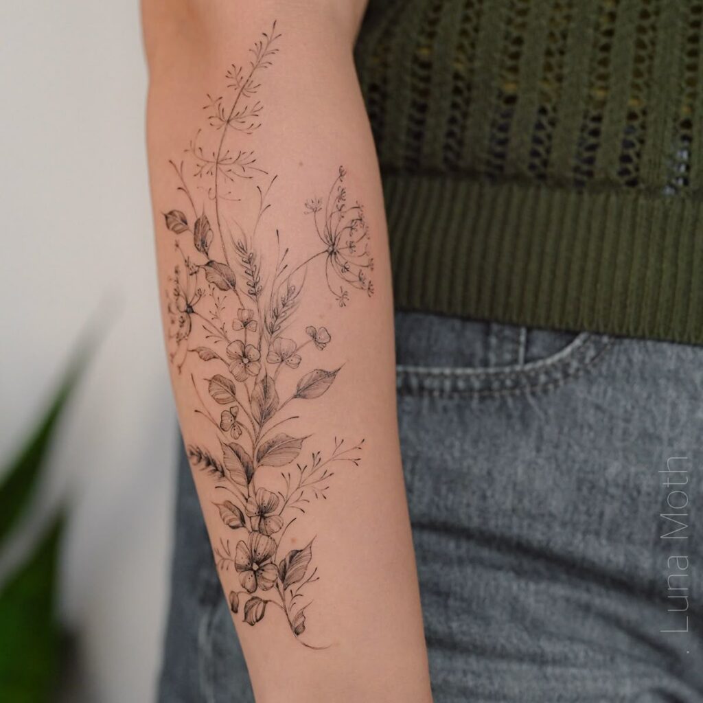 Delicate floral black ink tattoo on forearm, featuring intricate leaves and flowers, paired with a green sweater and denim jeans.